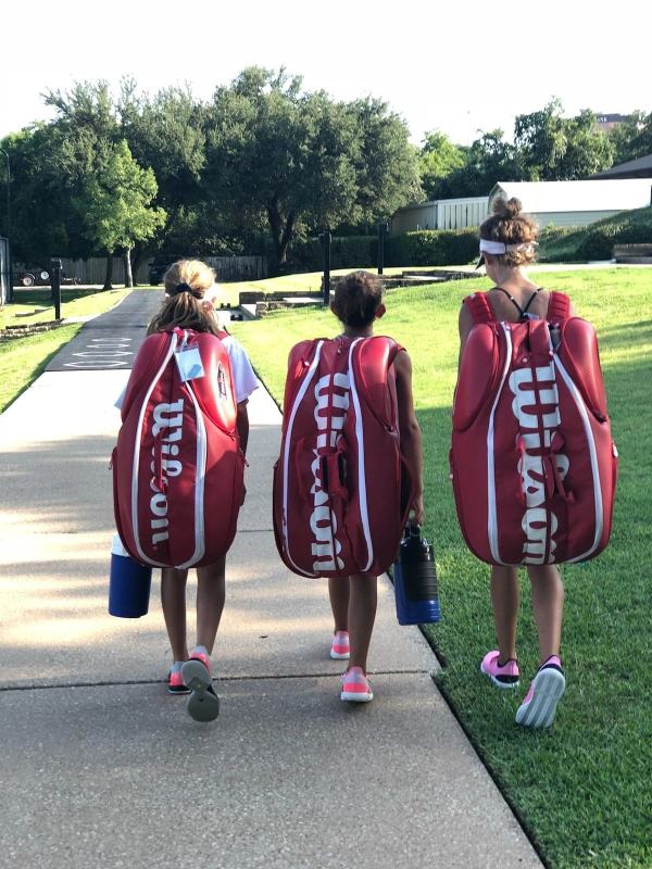 Three girls are walking down the sidewalk with tennis bags.